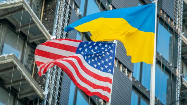 A Ukrainian flag and an American flag fly against a blue sky in front of a building